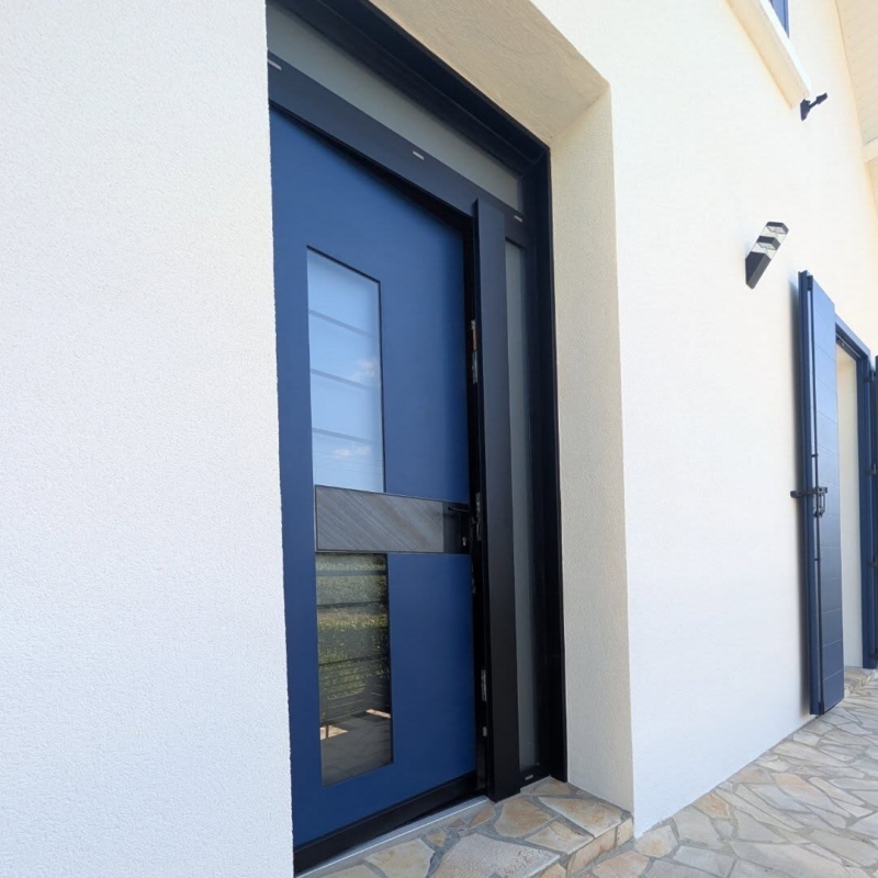 Vue en angle de la porte d'entrée aluminium Monalu Samouraï bleue et de son fixe vitré, façade enduite blanche
