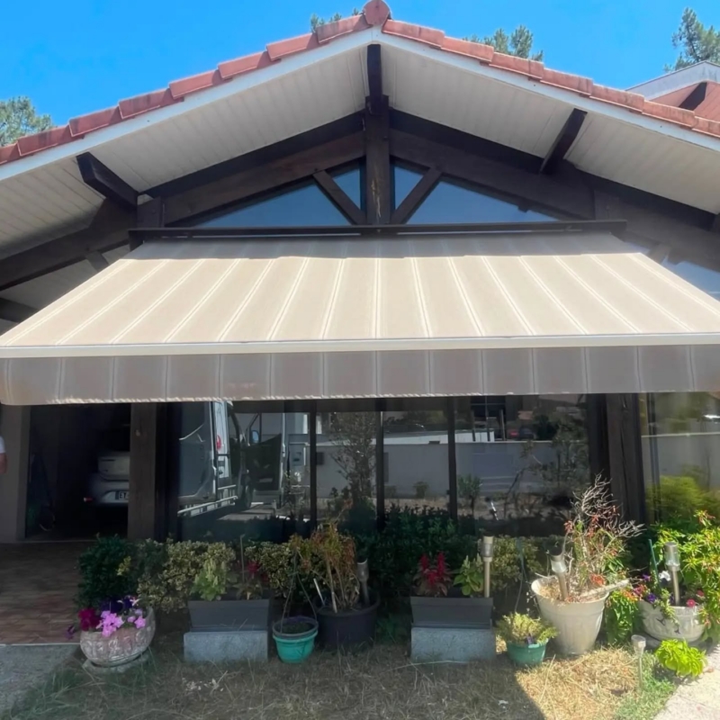 Store banne rénové avec toile neuve à rayures beige et sable, déployé sous le pignon d'une maison à charpente apparente à Castets