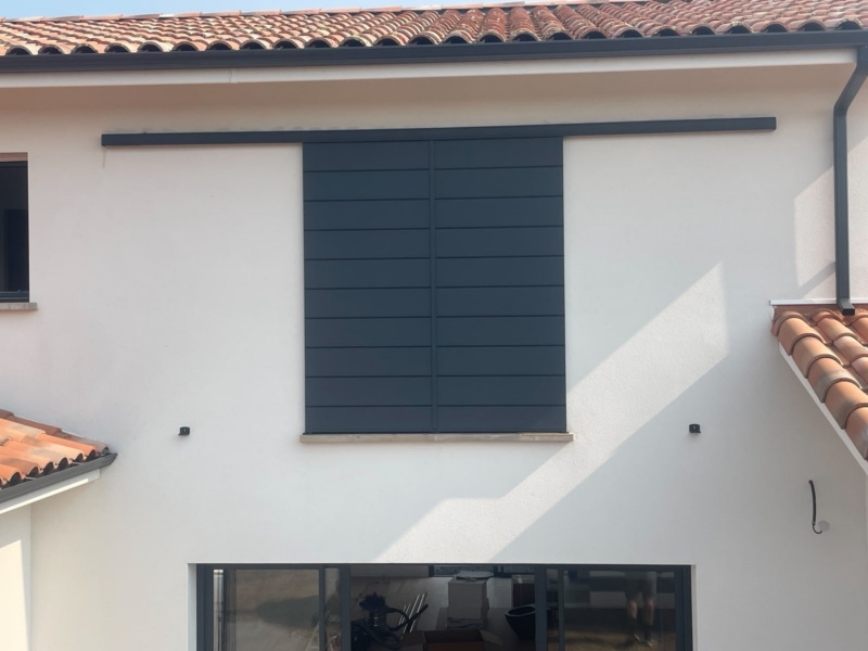 Volet coulissant aluminium gris anthracite à deux vantaux fermé devant la fenêtre de l'étage d'une maison blanche à Mimizan
