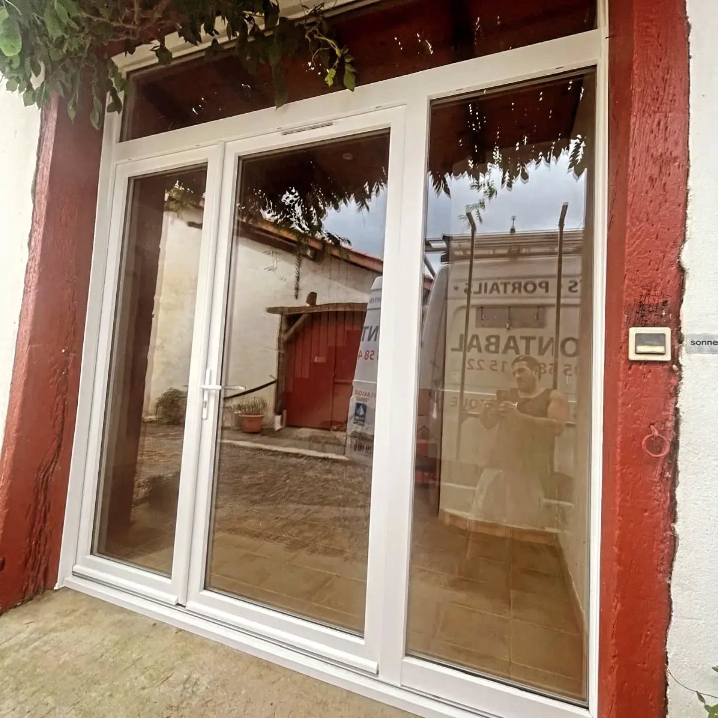 Ensemble vitré PVC blanc avec imposte posé entre des poutres peintes en rouge sur la façade d'un bâti traditionnel à Misson