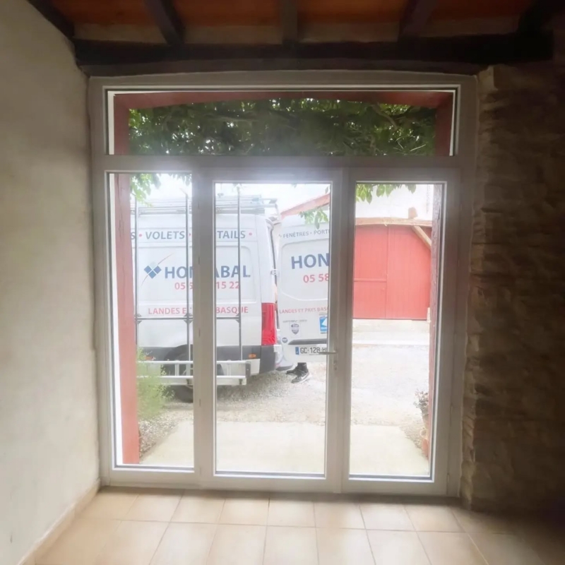 Vue intérieure d'une porte-fenêtre PVC avec imposte, mur de pierre et poutres au plafond, bâti ancien à Misson