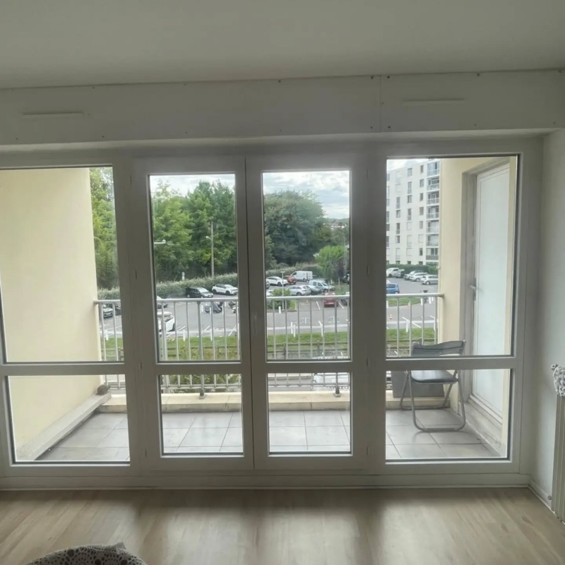 Vue intérieure d'une porte-fenêtre PVC blanche à deux vantaux entre deux châssis fixes, ouvrant sur un balcon à Anglet